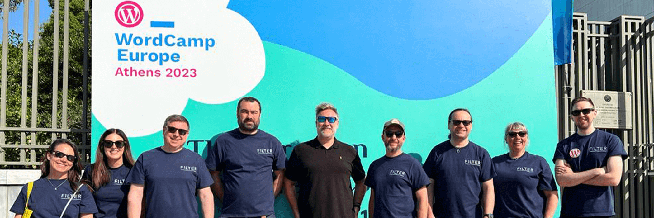 A group of eight people wearing matching navy shirts stand in front of a large blue and white sign for WordCamp Europe 2023 in Athens. They are outdoors, posing under clear skies, and smiling at the camera.