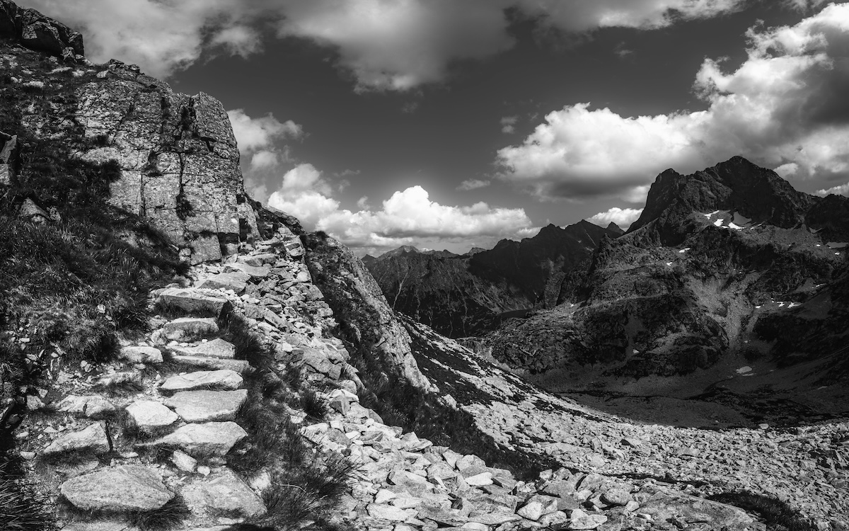 Rocky mountain path with stone steps, overlooking rugged peaks under a partly cloudy sky in black and white.